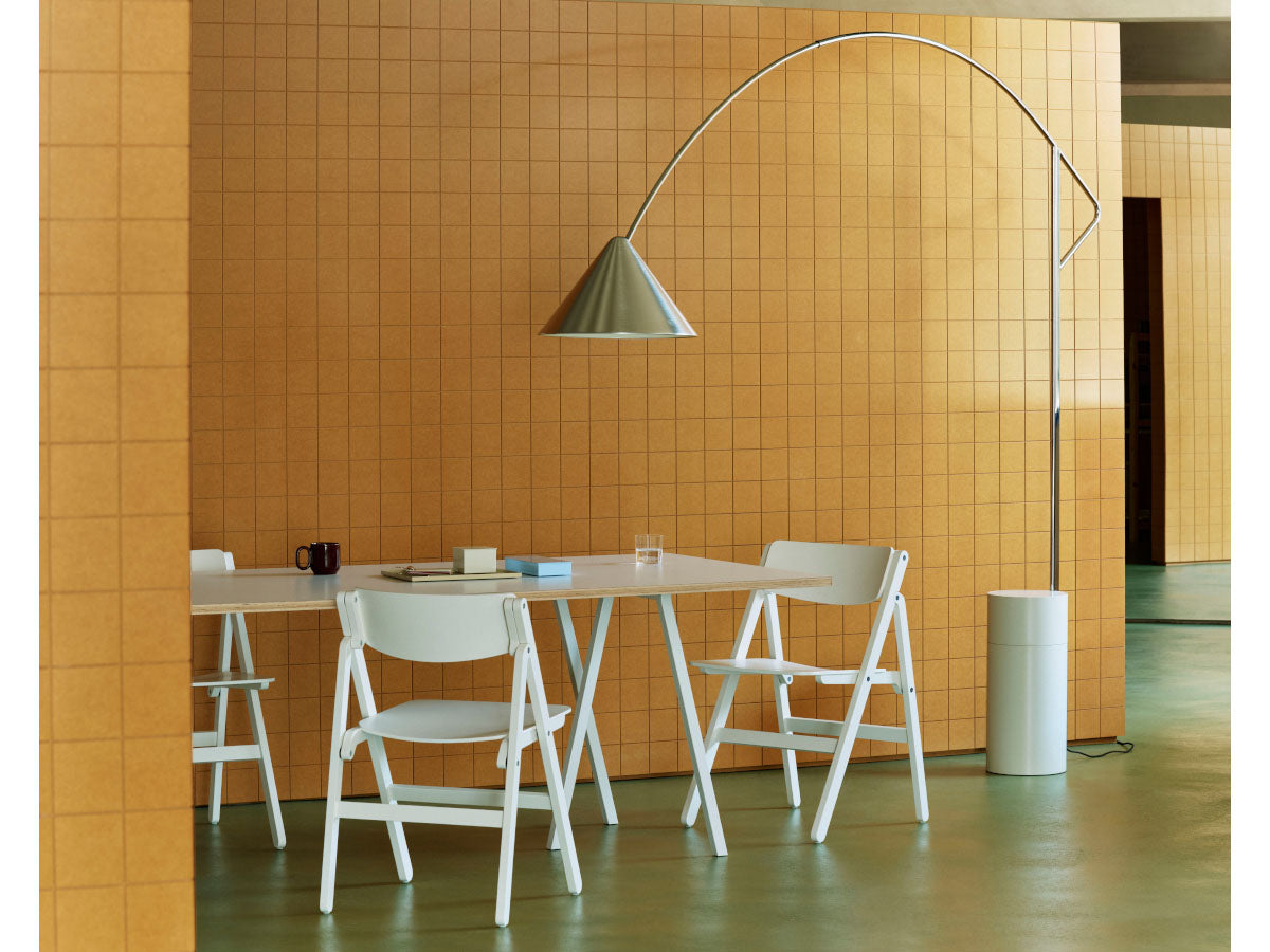 A modern interior scene with a white dining table and three white chairs set against a warm orange tiled wall, with a tall silver floor lamp and a white cylindrical side container.