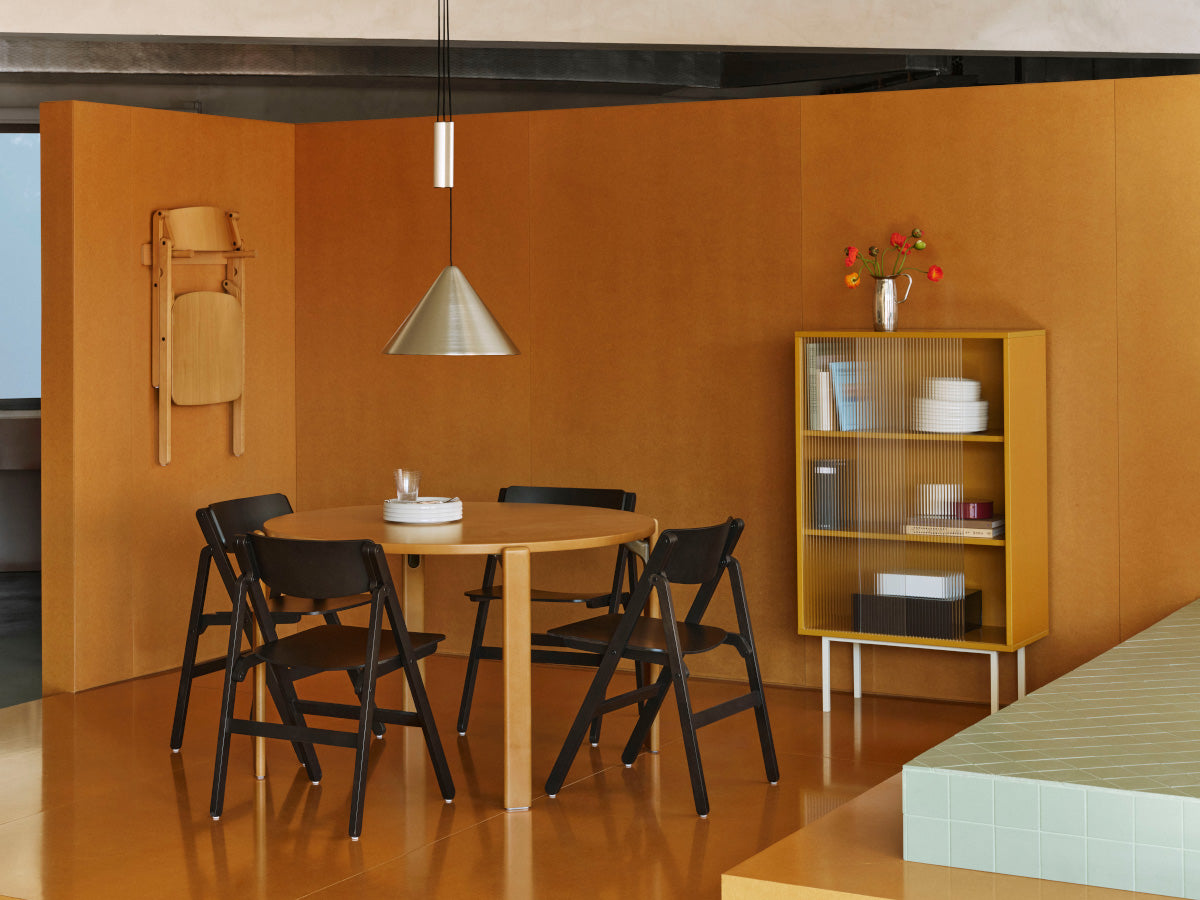 A modern dining area with a round light wood table surrounded by four black folding chairs, set against warm orange-brown walls. A silver cone pendant light hangs above, and a yellow shelving unit stands to the right holding books and decorative items.